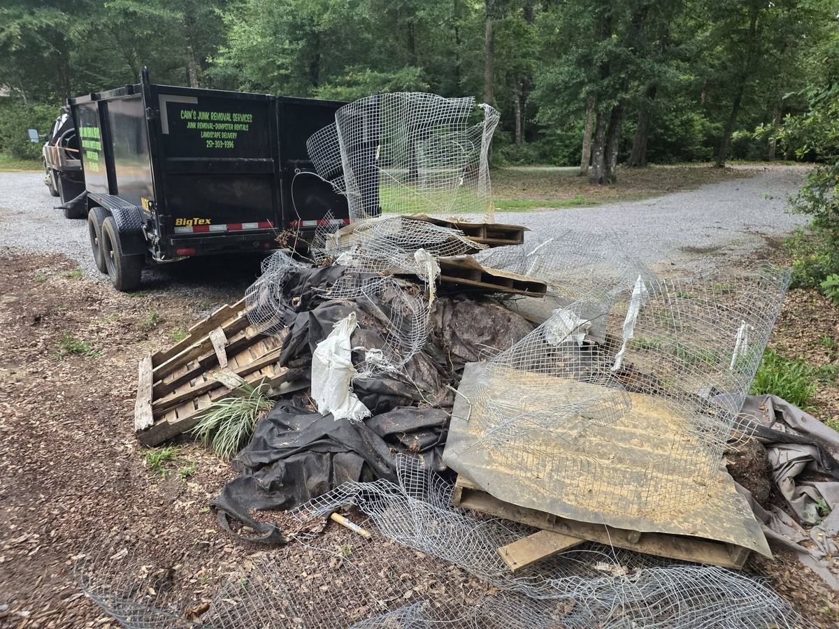 Pile of broken pallets, wire fencing and landscape fabric heaped on a gravel driveway beside Cain's black dump trailer.